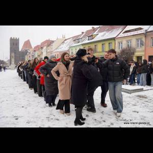 A line of people in winter attire stands on a snowy street, smiling and chatting with colorful buildings in the backdrop.