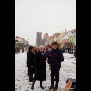 People stand in a snowy plaza, chatting, with colorful buildings and a tower in the background.