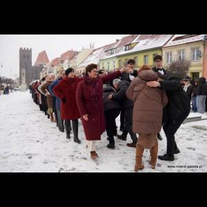 People line up in the snow while a woman guides them, colorful buildings in the background.