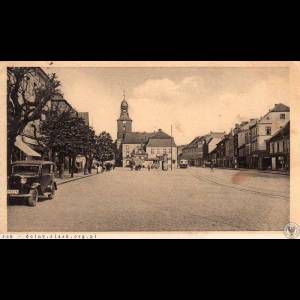 A vintage town square with a clock tower, lined by trees and buildings, featuring classic cars and people.