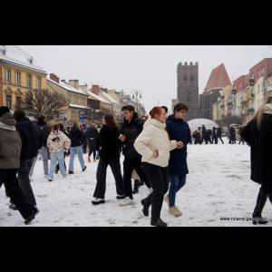 A snowy public square filled with people walking, dressed in winter attire, with colorful buildings in the background.