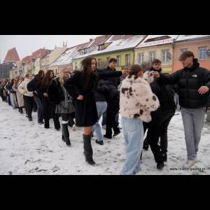 A line of people dressed in winter attire walking on snow, engaging in a fun activity in a colorful urban setting.
