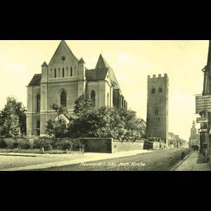 Historic street view featuring a Catholic church and a tall tower, surrounded by trees and buildings.