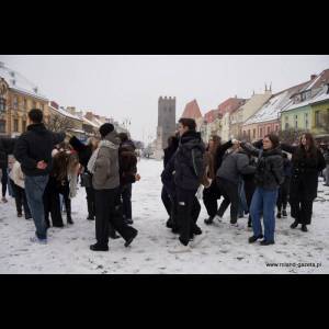 A snowy square filled with people dressed in winter clothes, interacting near colorful buildings and a tower.