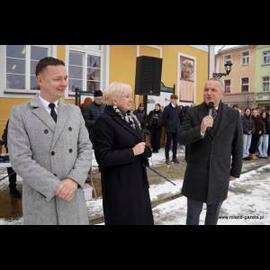 Three speakers stand outdoors, with an audience in the background, on a snowy day near a colorful building.