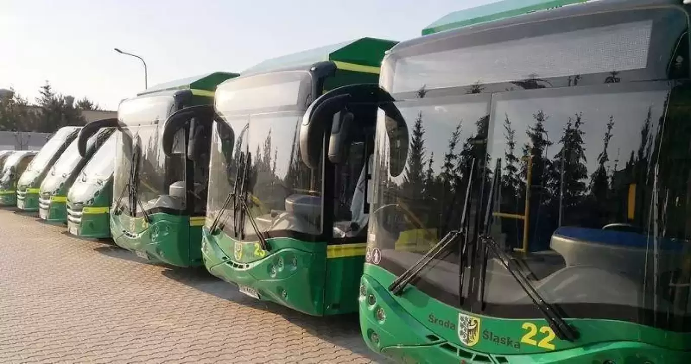 Rząd zielonych, nowoczesnych autobusów miejskich ustawionych na placu w Środzie Śląskiej; najbliższy oznaczony numerem 22, w tle drzewa i jasne światło dnia.