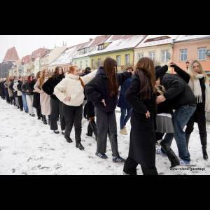 A line of people in winter clothing, posing playfully in the snow along a street with colorful buildings.