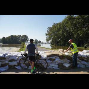 A cyclist observes floodwaters alongside a sandbag barrier, while another person in a vest works nearby.