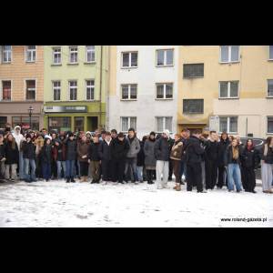 A large group of students stands together in a snowy area, dressed in winter coats, near colorful buildings.