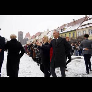 People dancing in a snowy square, surrounded by colorful buildings and a crowd watching.