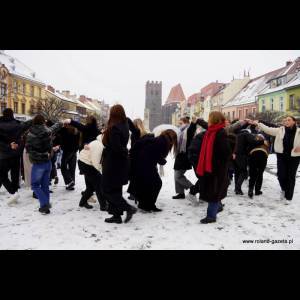 A snowy square with a crowd dancing or moving, colorful buildings, and a tower in the background.