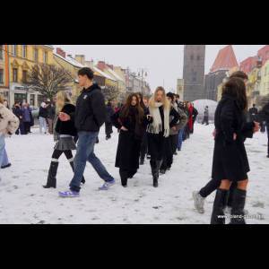 A snowy scene with people walking in line, wearing winter clothing, amidst colorful buildings and a clock tower.