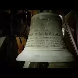 A close-up of a large, ornate bell with Latin inscriptions, hanging in a wooden structure.