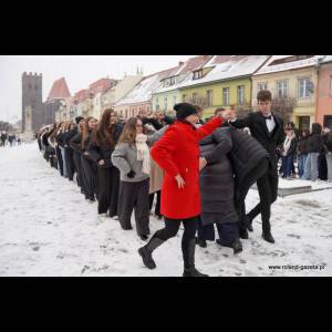 A group of people in winter clothes forms a line while a woman in red leads in a snowy urban setting.
