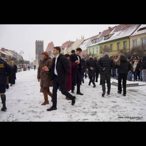 A snowy street scene with people walking, some dressed formally, and colorful buildings in the background.