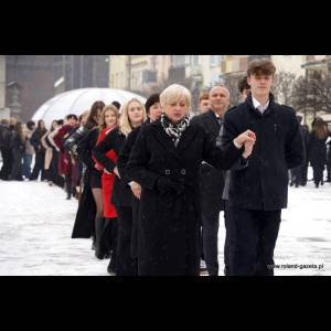 A group of people in formal attire walking through snow, led by a woman in a black coat and scarf.