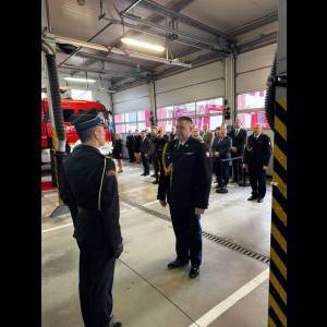 A formal event in a fire station with officials and guests, featuring military attire and a fire truck in the background.