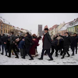 A group of people gathering in snow, some dressed formally, in a historic town square with buildings and towers.
