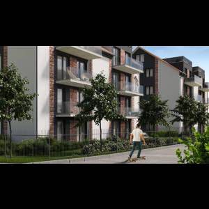 A person skateboards past modern apartment buildings with greenery and fences around the area.