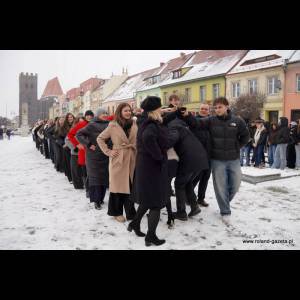 A line of people in winter attire stands on snowy ground, with colorful buildings and a tower in the background.